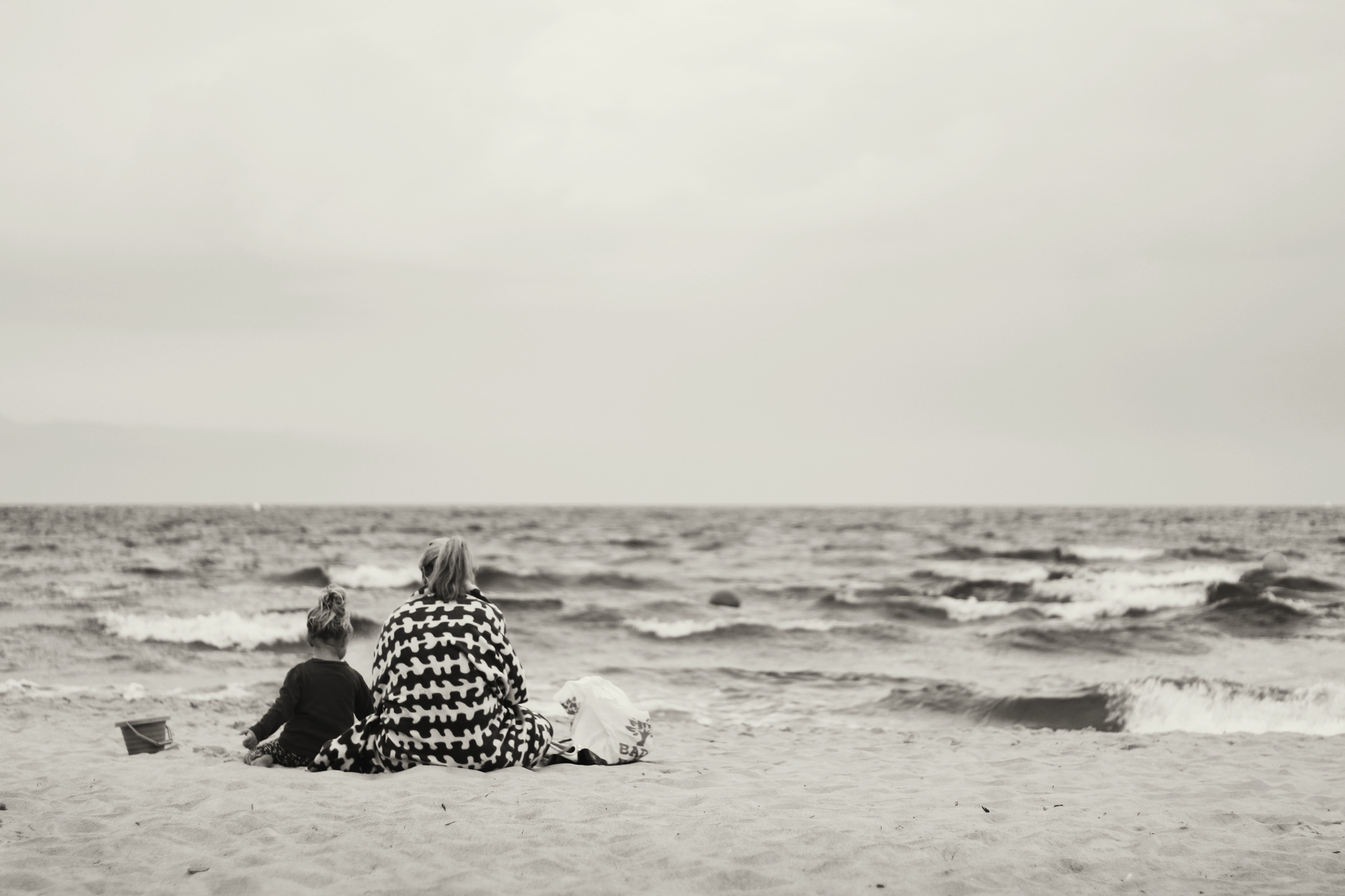 Woman And Child Sitting On Sand Near Body Of Water